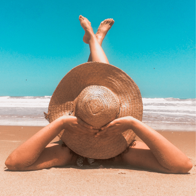 Imagem de uma mulher com chapéu de palha, deitada na areia com as pernas para cima na praia